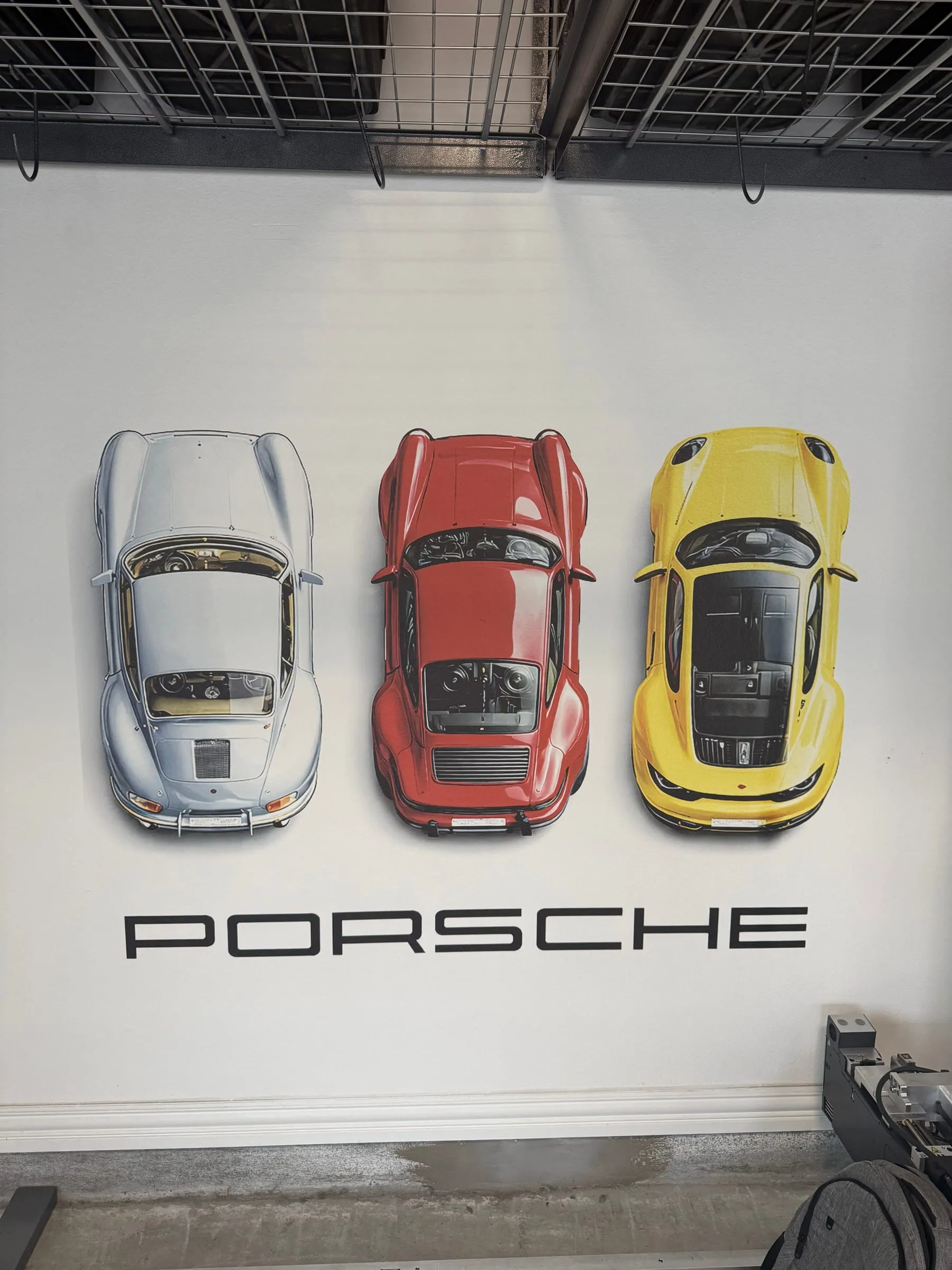 Three Porsche cars seen from above, in silver, red, and yellow, over a PORSCHE wordmark on a garage wall